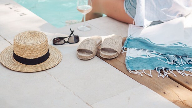 A poolside setting with a person sitting by the pool, a hat, sunglasses, and a towel on the table.