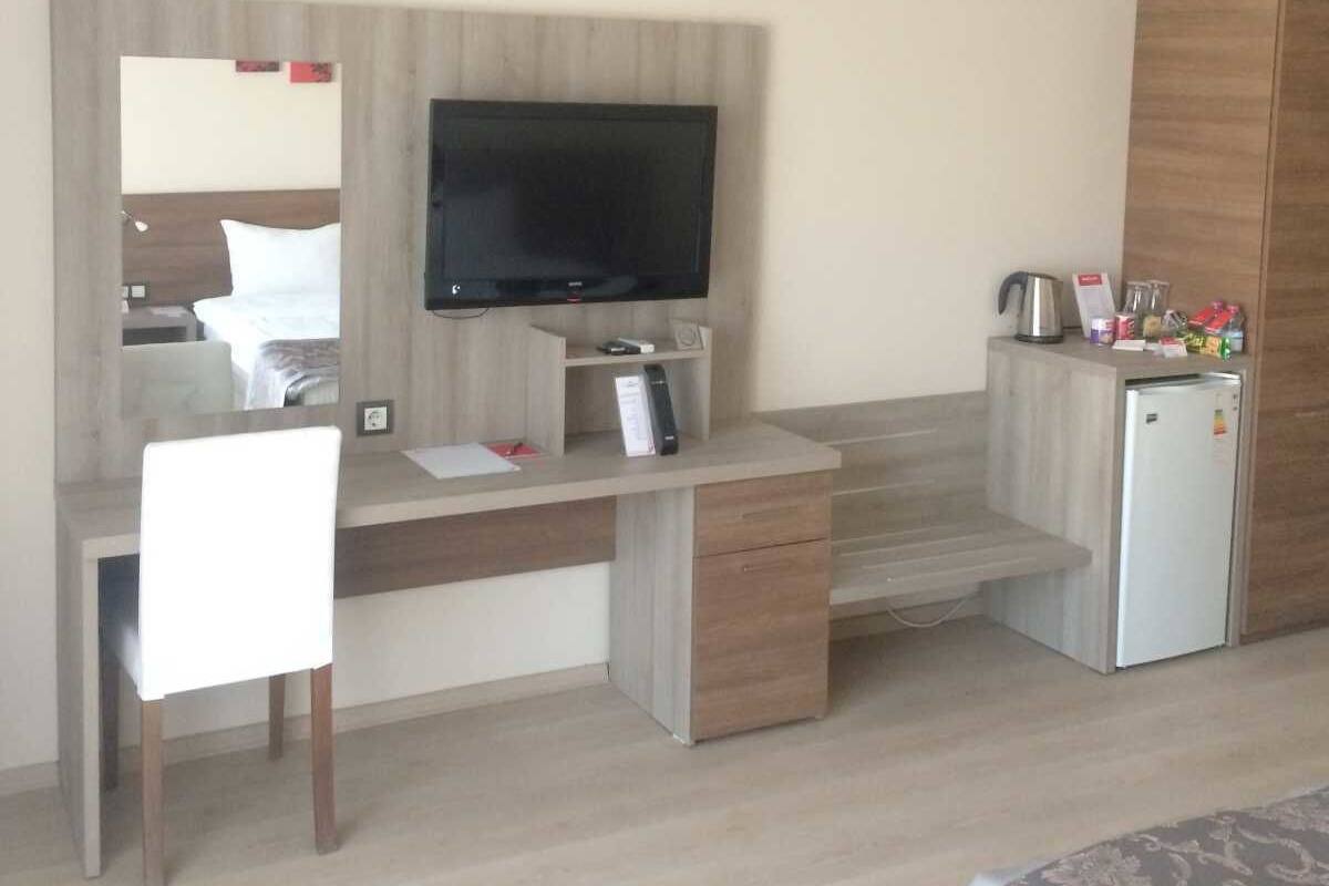 A hotel room with a wooden desk, a television, a mini-fridge, and a kitchenette.