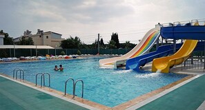 A swimming pool with multiple slides, including a rainbow-colored one, surrounded by lounge chairs and umbrellas. - Geyikli Aqua Otel (Ezine)