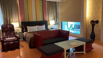 A hotel room with a bed, a red sofa, a small table, a leather armchair, a nightstand with a lamp, and a window with curtains.