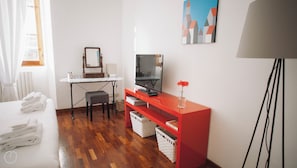 A hotel room with a wooden floor, a desk with a mirror, a television, a red console table, a lamp, and a window with curtains.