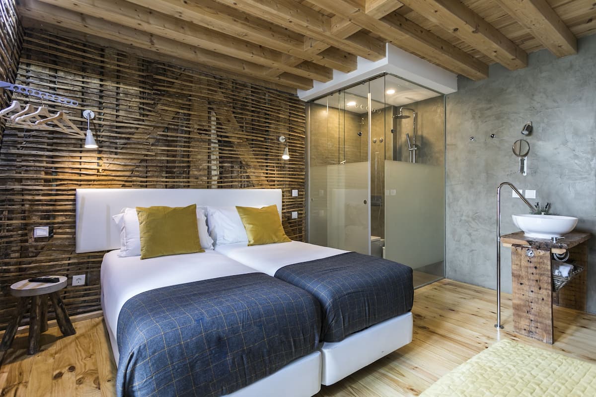 Un dormitorio moderno con una cama grande, vigas de techo de madera, un box de ducha de vidrio y un baño pequeño con lavamanos y espejo.