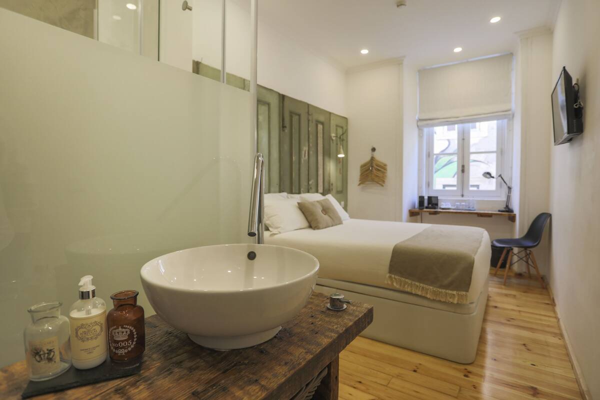 Un baño moderno con un lavamanos grande de color blanco, un mueble de tocador de madera y una cama con una manta color beige.