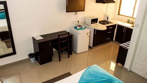 A compact hotel room with a kitchenette, a desk, a chair, a small refrigerator, and a bed with a blue cover. - Laha Hotel (Maun)