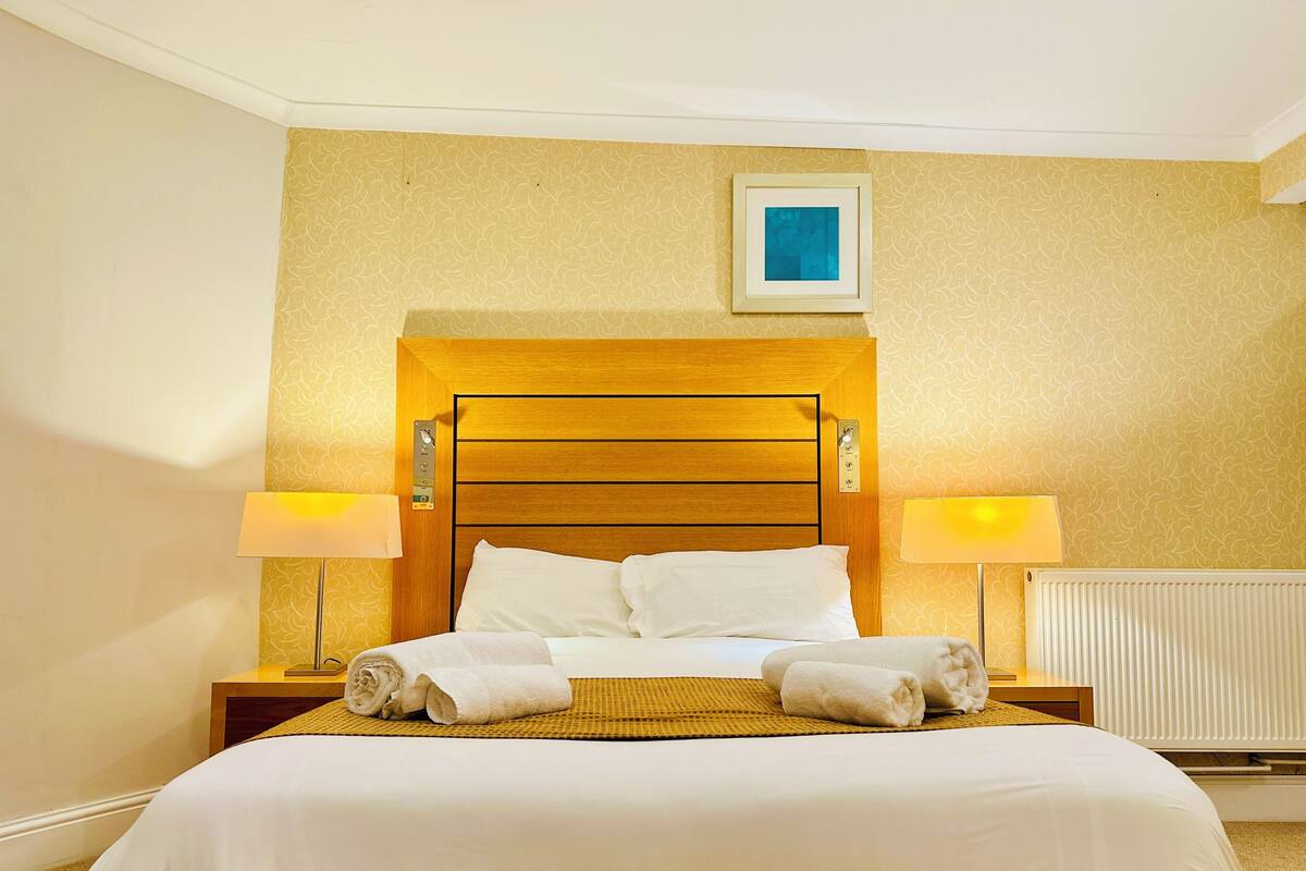 A hotel room with a wooden headboard, two bedside lamps, a bed with white linens, and a framed picture on the wall.