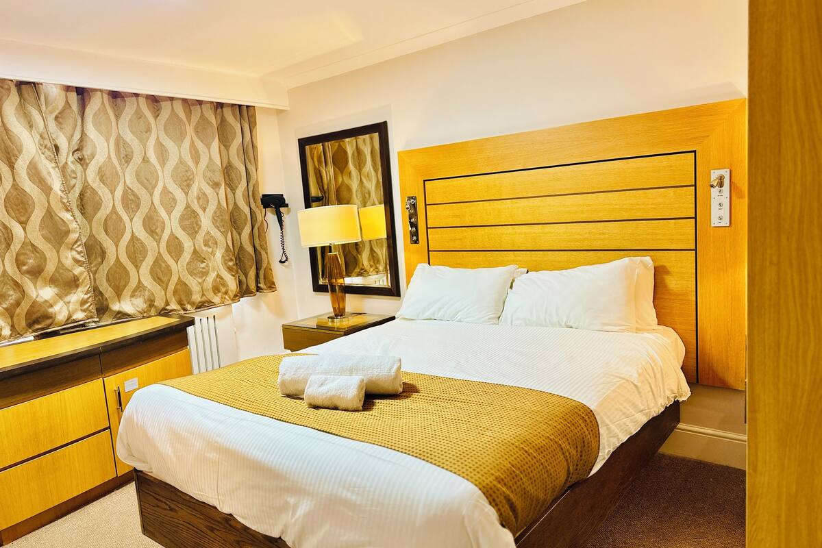 A hotel room with a large bed, wooden headboard, a bedside table with a lamp, a mirror, and a patterned curtain.