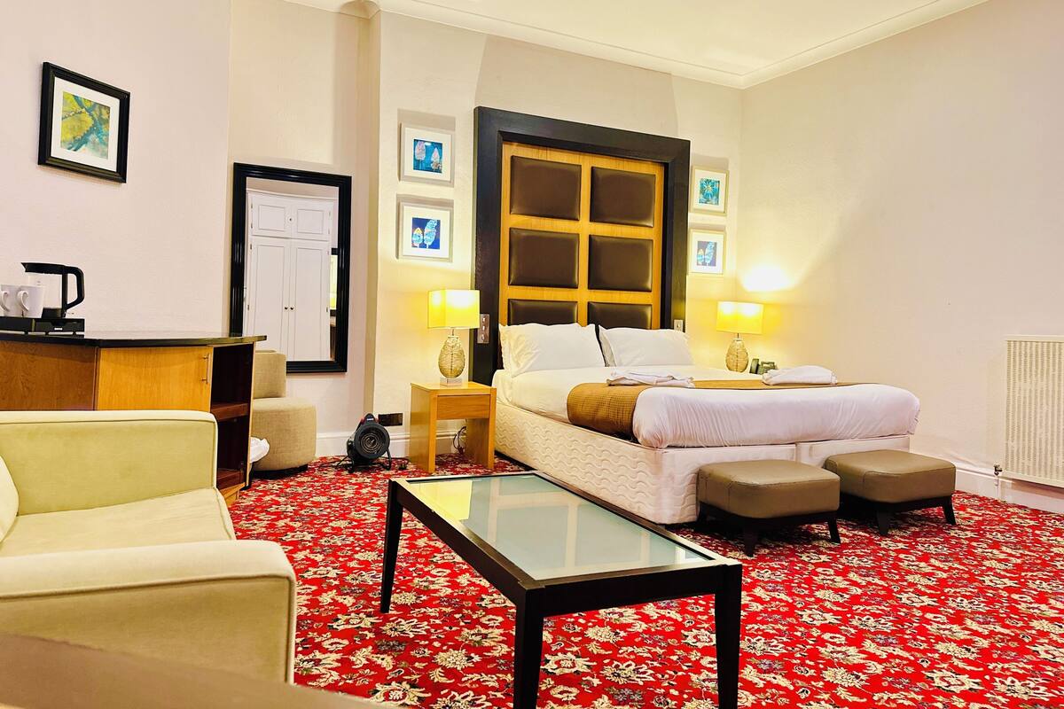 A hotel room with a bed, two bedside lamps, a desk with a coffee maker, a chair, and a red floral carpet.