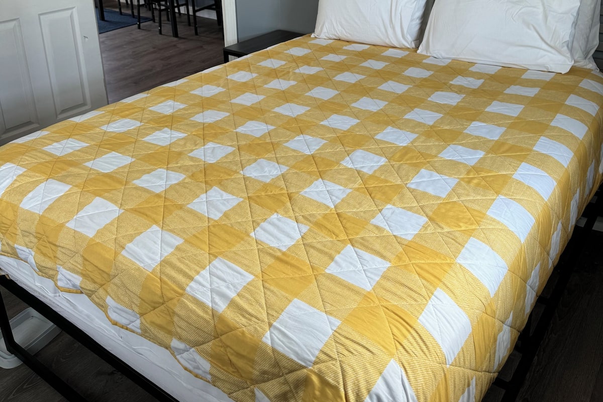 Una cama con una colcha a cuadros amarillo y blanco, una puerta blanca, y una pequeña mesa con sillas al fondo.