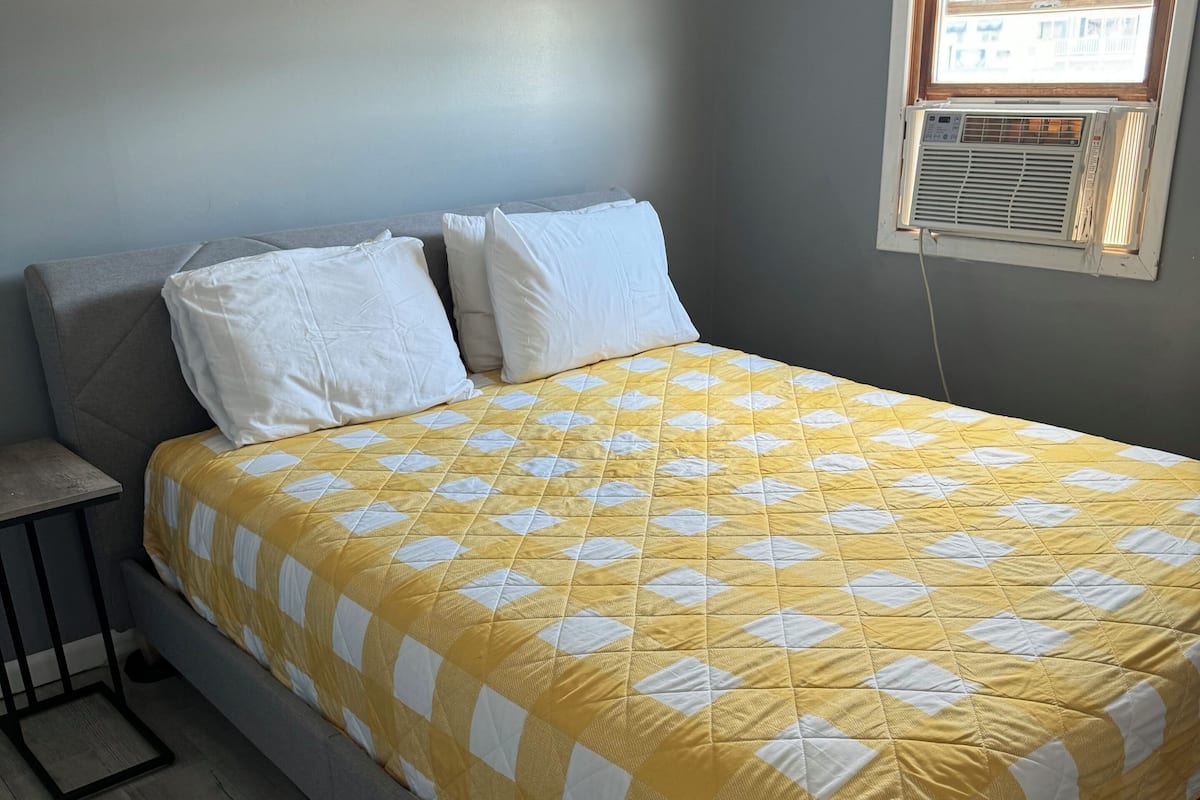 Un dormitorio con cama, almohadas, una mesita de noche y una ventana con aire acondicionado.