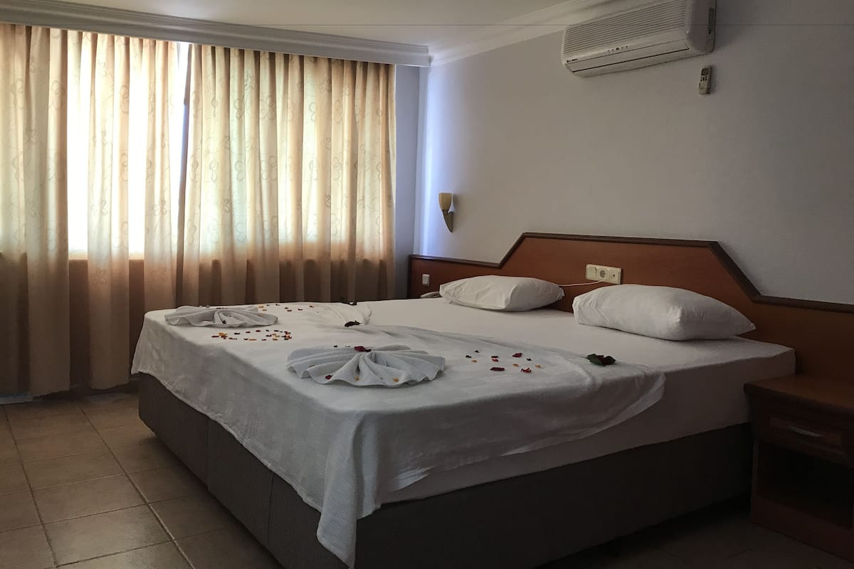 A hotel room with a large bed, a bedside table, an air conditioning unit, and a window with curtains.