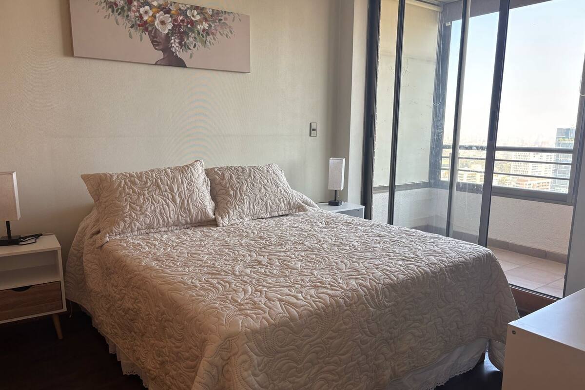 Una cama bien hecha con una colcha de diseño floral, una mesita de noche con lámpara y un ventanal con vista a la ciudad.