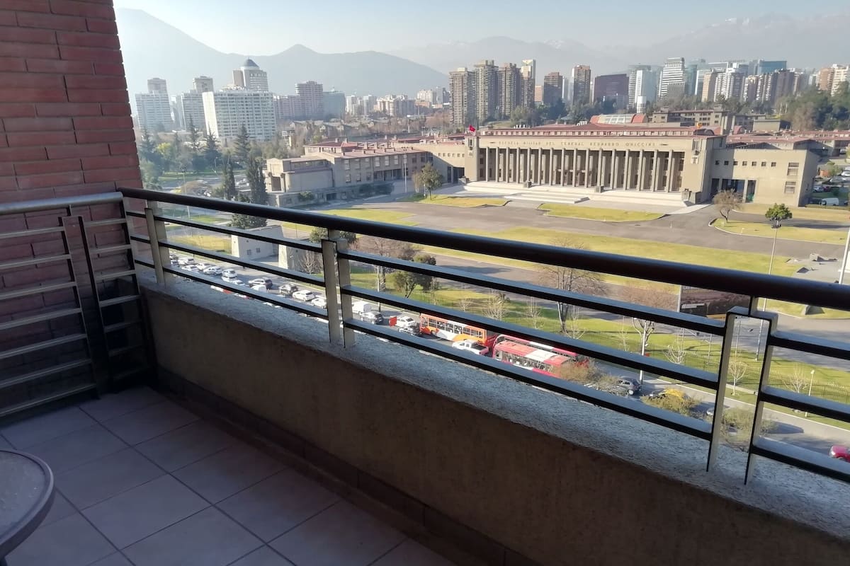 Un balcón con vista a un paisaje urbano, que incluye un edificio grande con columnas y un estacionamiento.