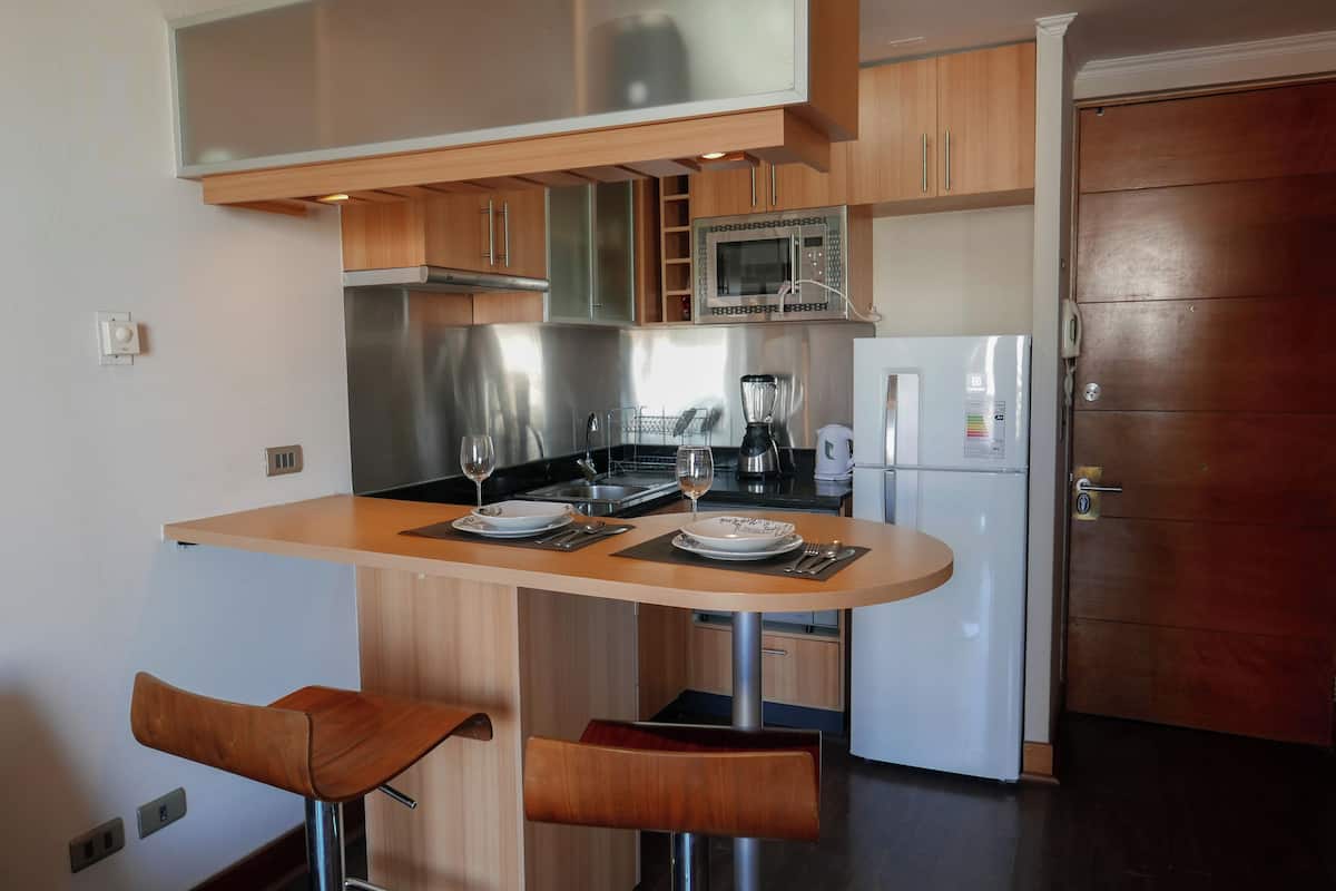 Cocina compacta con microondas, estufa y refrigerador. Hay dos taburetes de madera para bar y una pequeña isla con pileta.