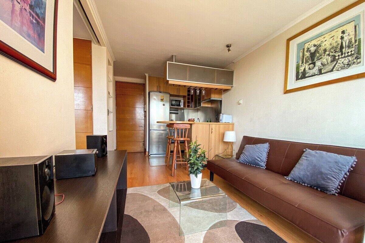 Una sala de estar moderna con un sofá marrón, una mesa de centro y un televisor. Se observa una cocina al fondo con un comedor.