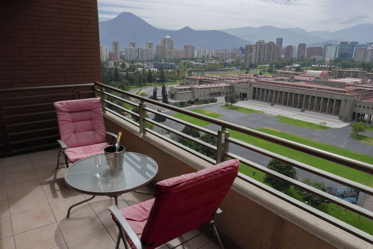 Un balcón con dos sillas rosas y una mesa de vidrio, con vista a un paisaje urbano que incluye edificios y uno con techo rojo.
