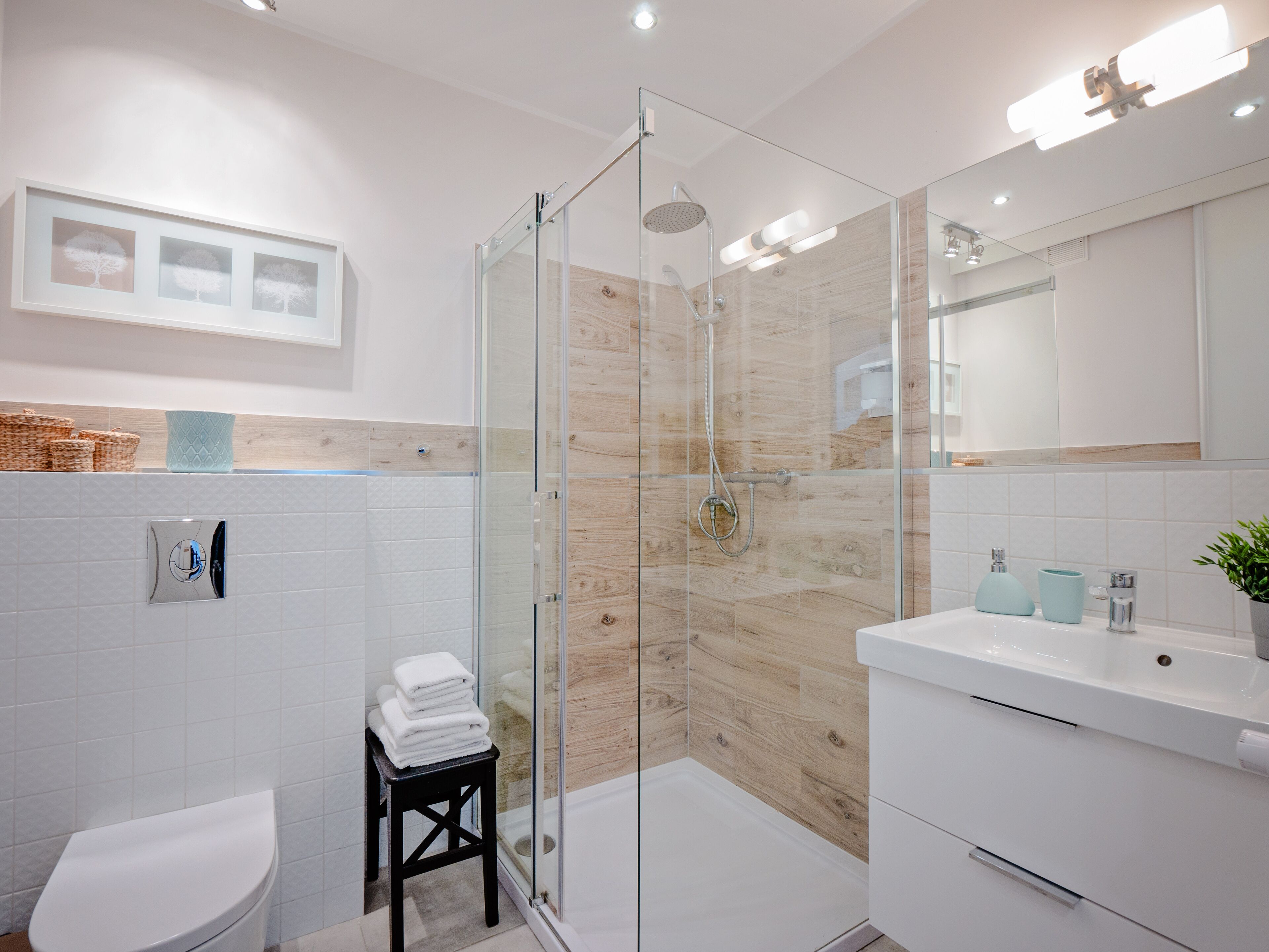 A modern bathroom with a glass-enclosed shower, white tiles, a wooden accent wall, a white sink, and a small stool with towels.