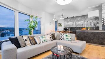 A modern living room with a grey sectional sofa, a round coffee table with candles, and a large window offering a view of the city.