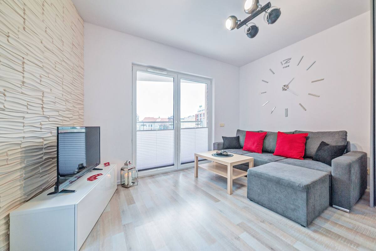 Un salon moderne avec un téléviseur à écran plat, un canapé gris agrémenté de coussins rouges, une table basse en bois et une grande horloge murale.