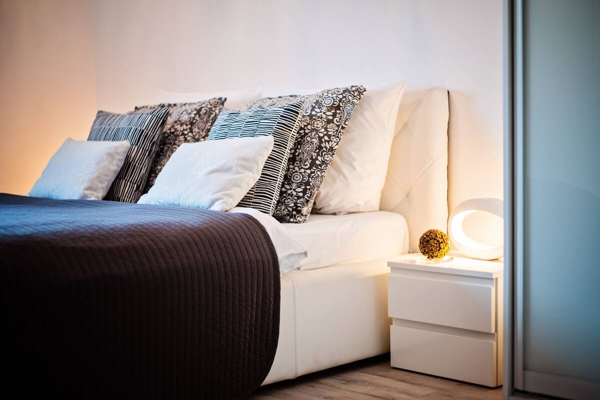 Un lit bien fait, recouvert d’une couvre-lit marron foncé et agrémenté de plusieurs coussins, une table de chevet avec une lampe et un petit objet décoratif.