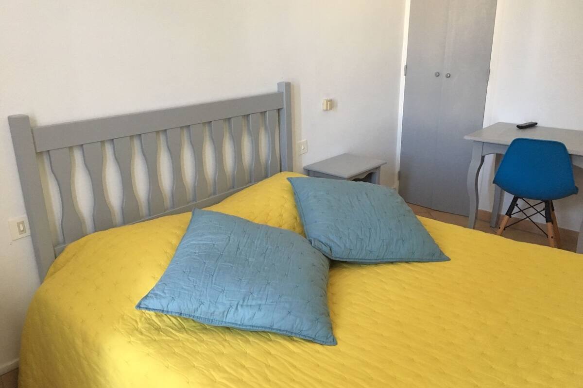 A bed with a yellow bedspread and two pillows, a grey headboard, a small bedside table, and a blue chair.
