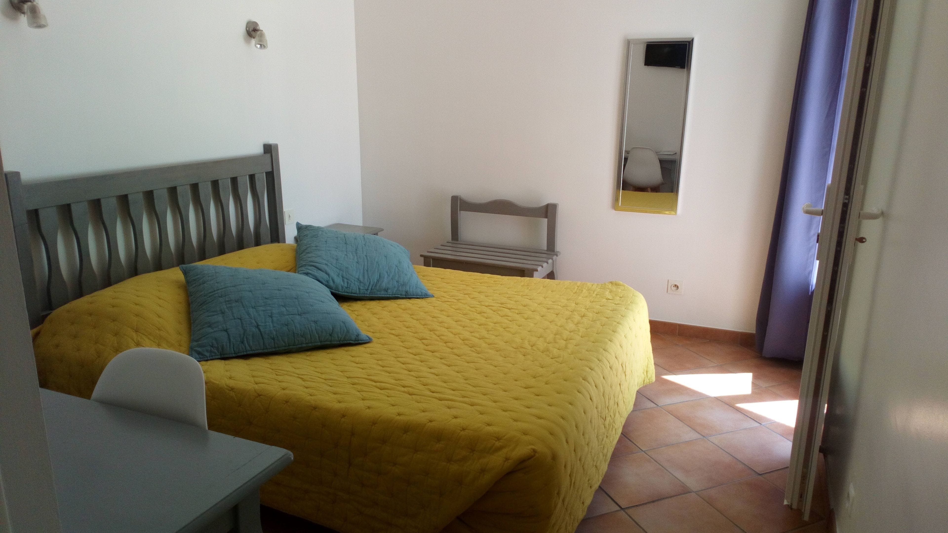 A bedroom with a bed, two pillows, a bedside table, a chair, a small table, a mirror, and a door.