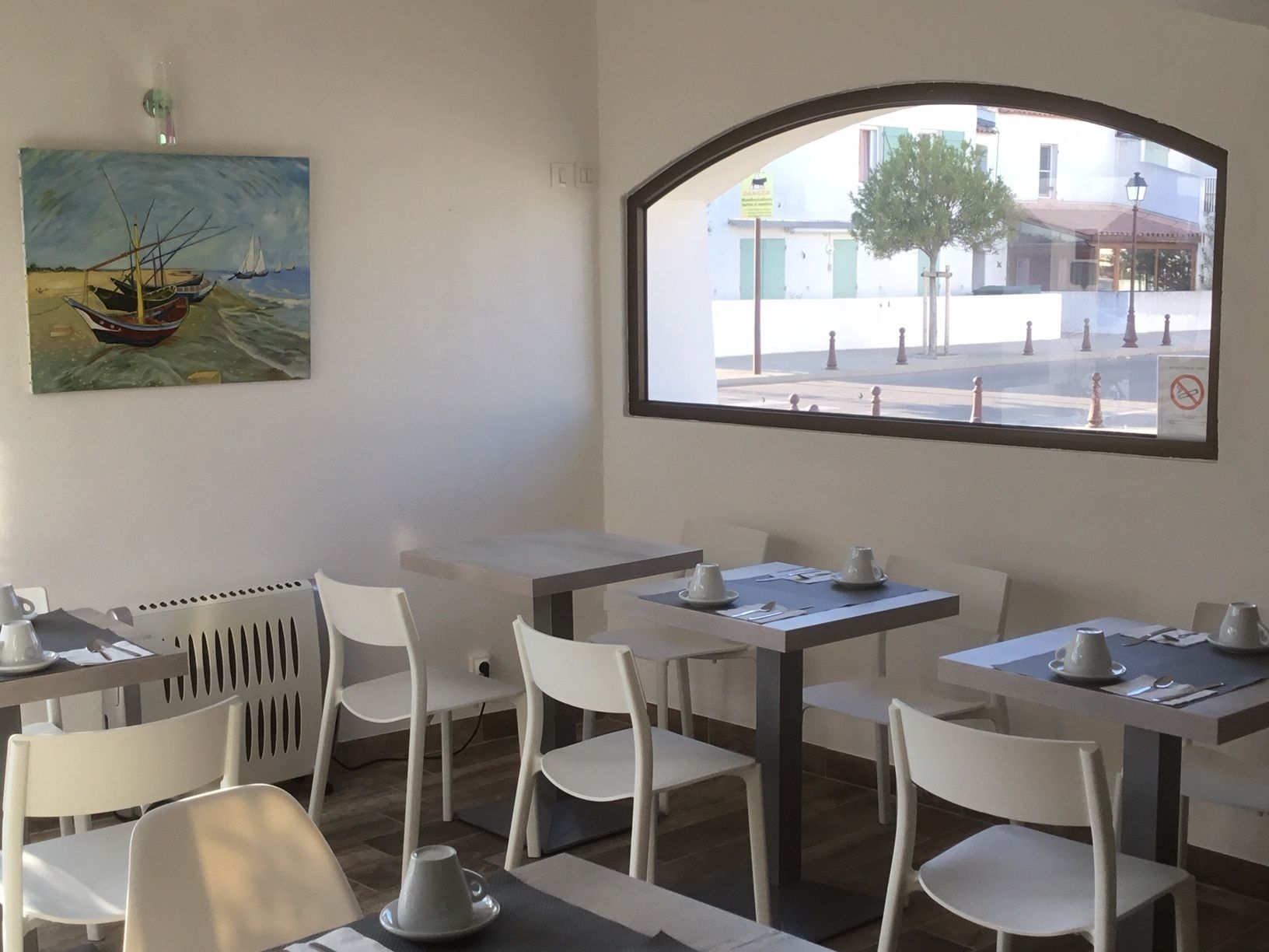 Un comedor con mesas y sillas blancas, un ventanal con vista a la calle y un cuadro de un barco en la pared.
