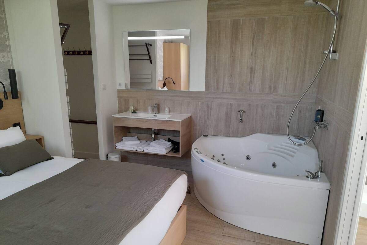 A modern bathroom with a large bathtub, a vanity with a mirror, and a bed with a gray headboard.
