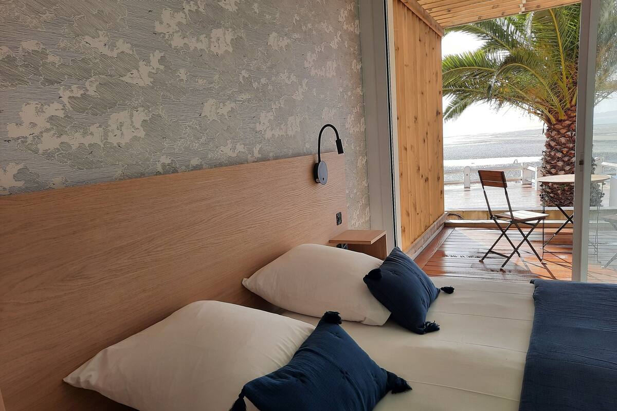 A bedroom with a wooden headboard, two pillows, a bedside table, and a view of the beach through large windows.