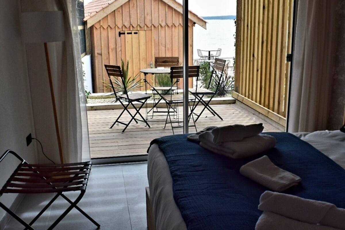 A bedroom with a bed, a chair, a table, and a view of a wooden building and water outside.