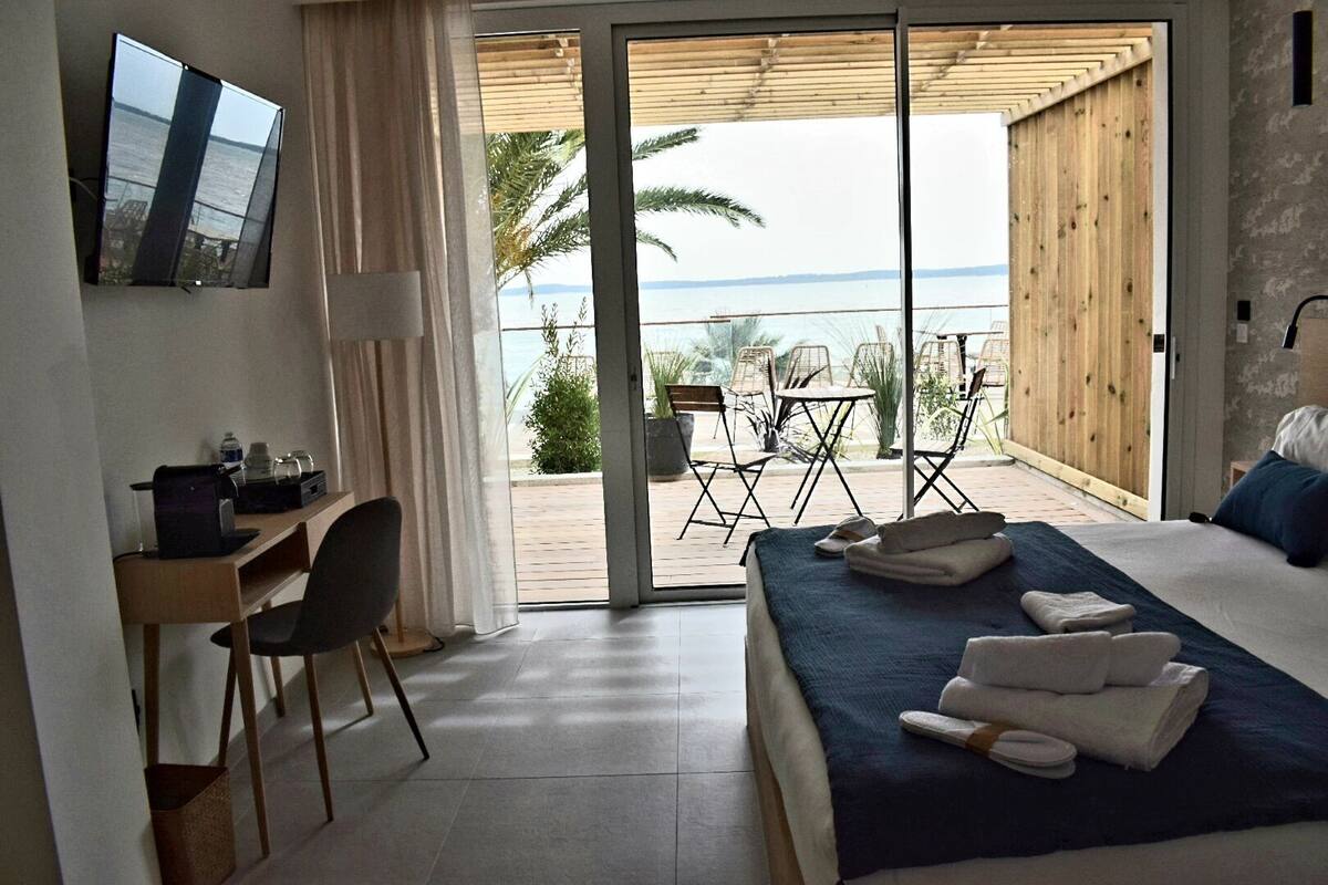 A hotel room with a bed, a desk, a chair, and a view of the sea through a sliding glass door.