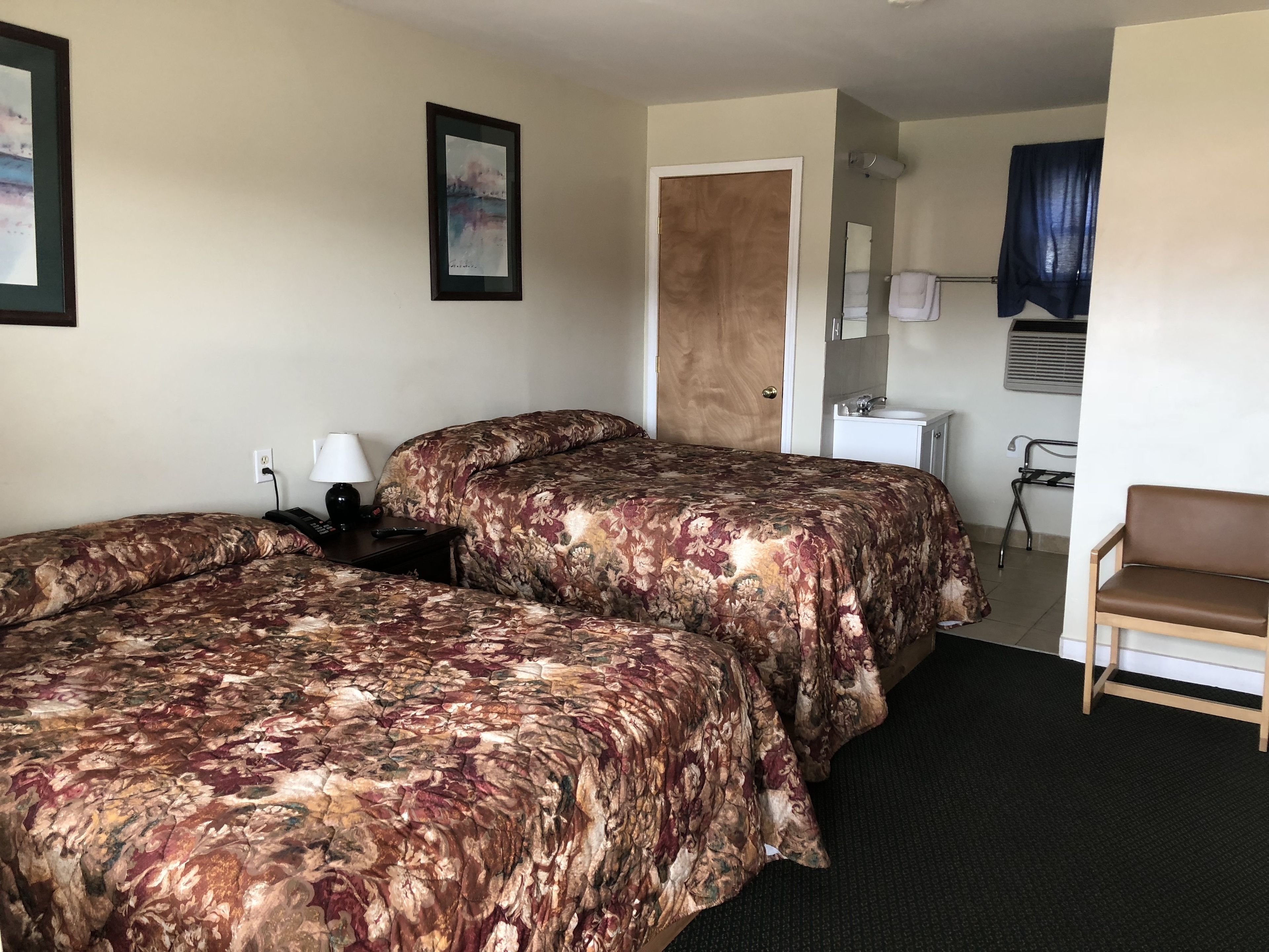 A hotel room with two beds, a desk, a chair, a bathroom, and a painting on the wall.