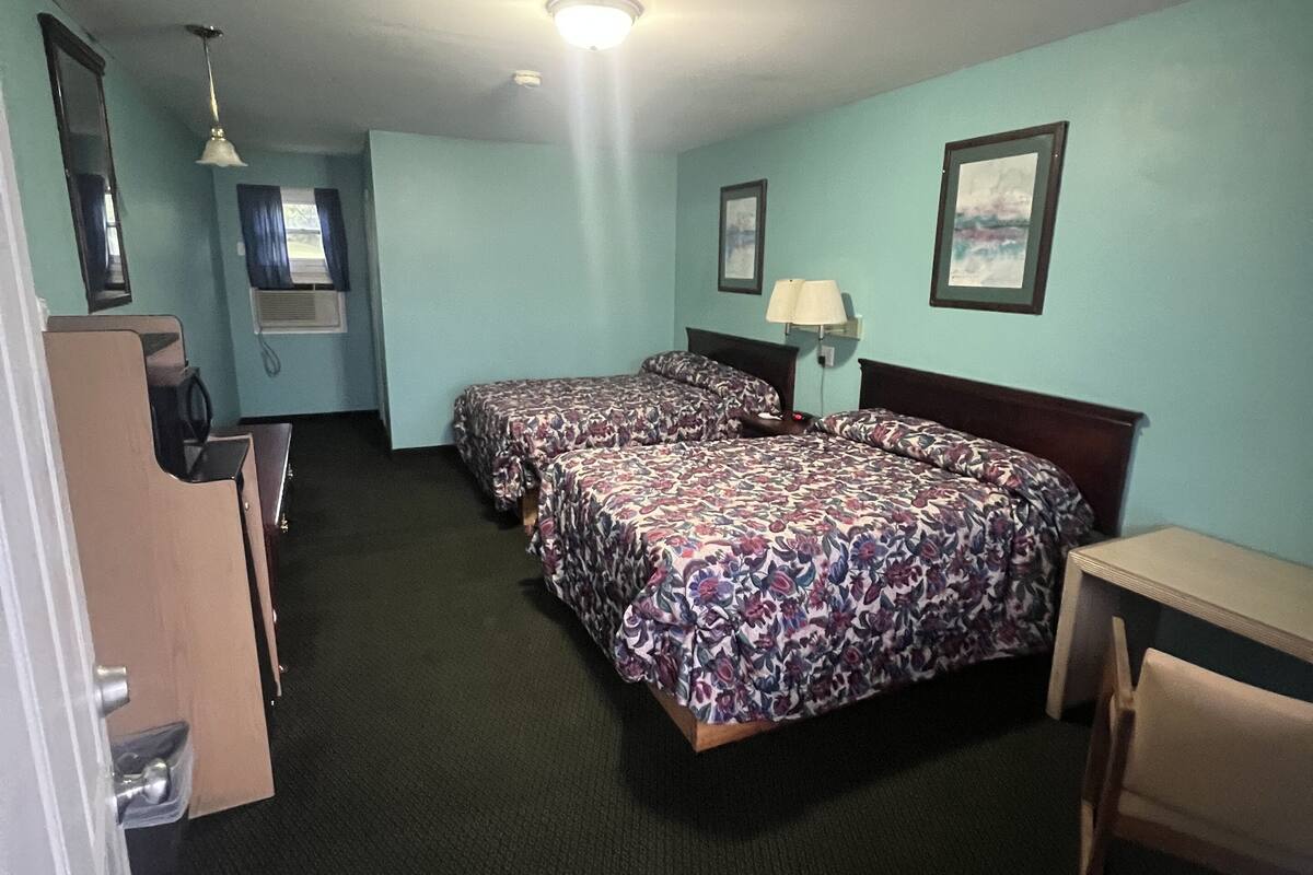 A hotel room with two beds, a desk, a chair, a lamp, a mirror, and a window with curtains.