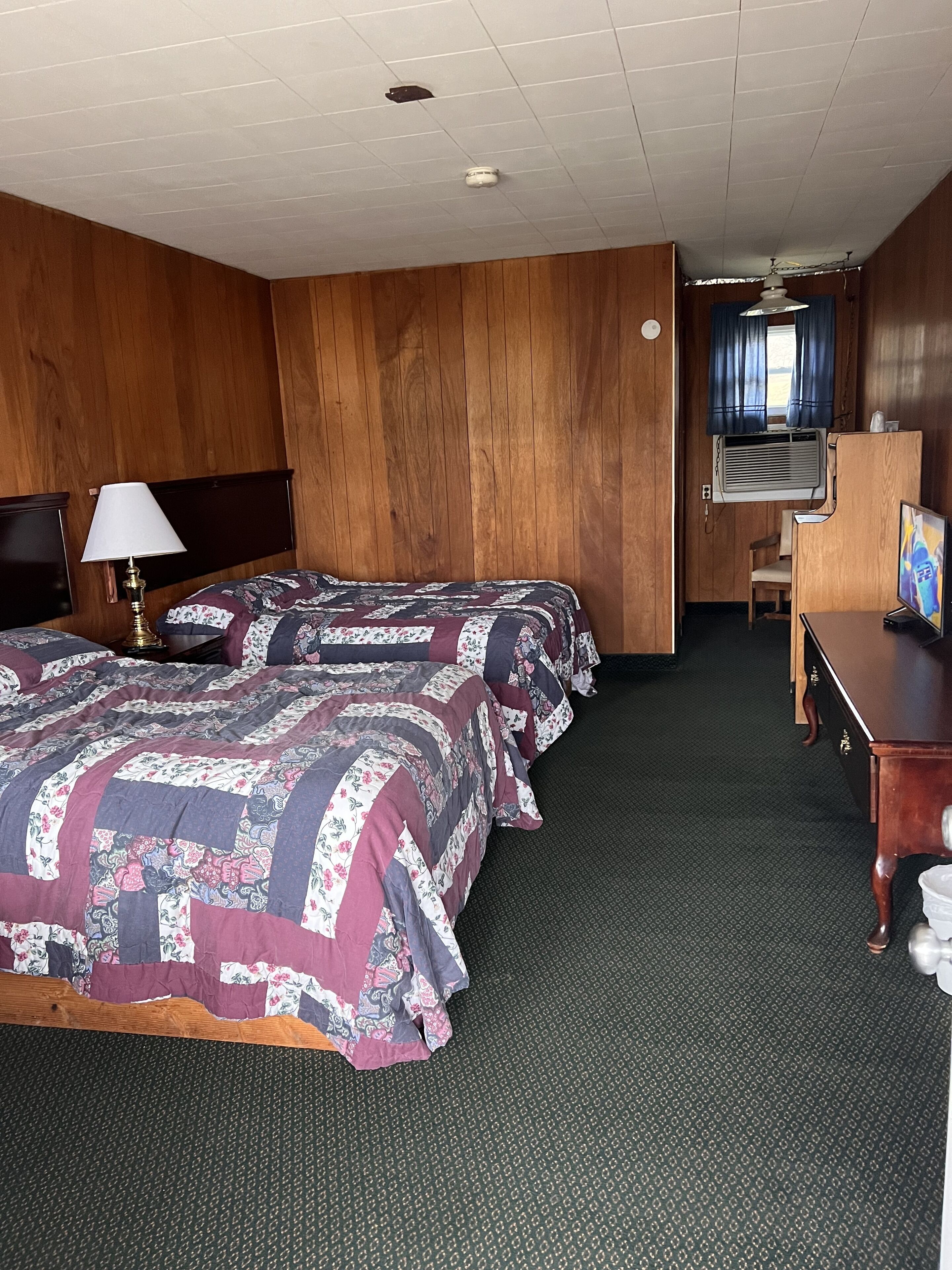 A hotel room with two beds, a desk, a TV, and an air conditioning unit.