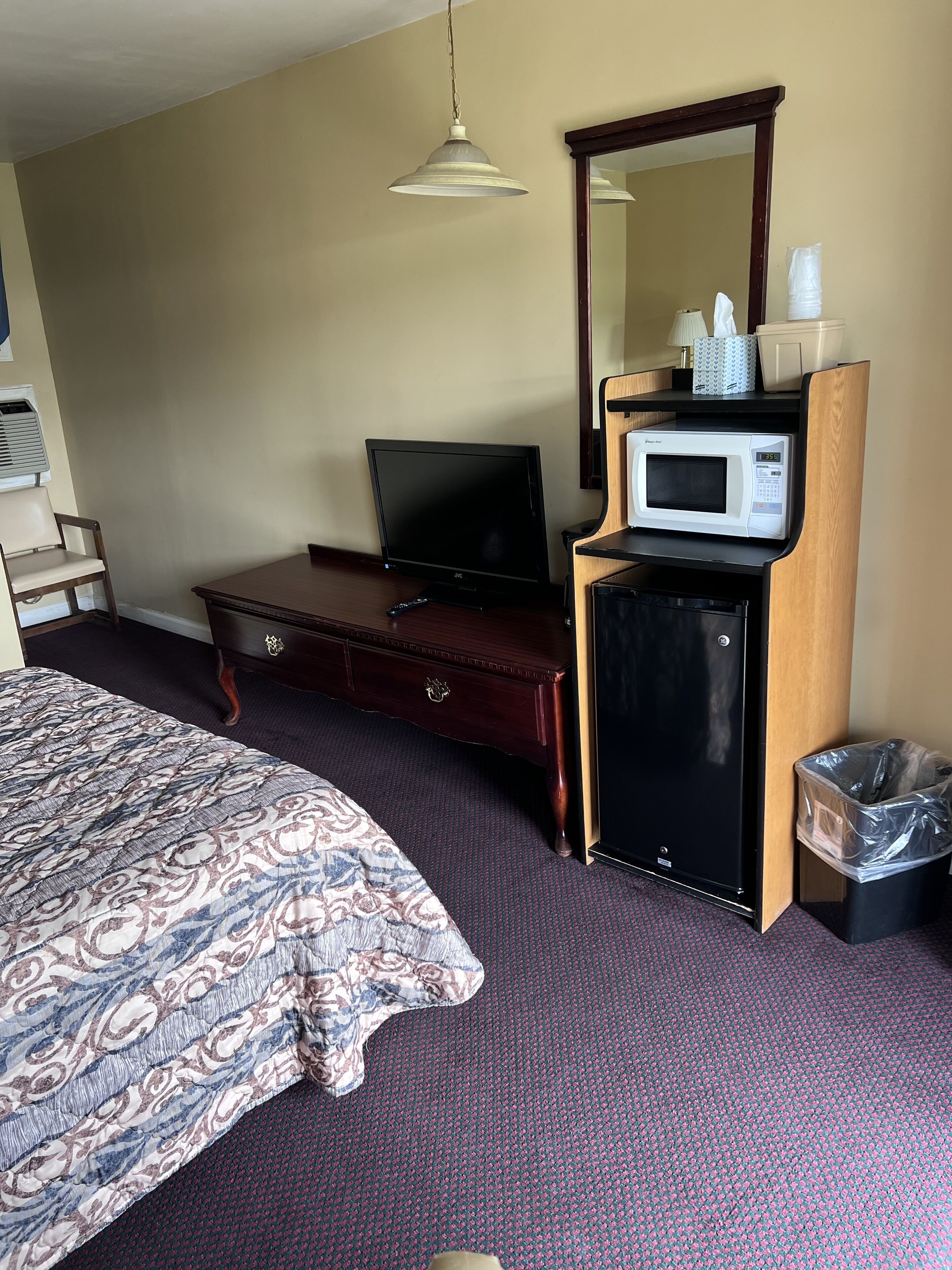 A hotel room with a bed, a dresser, a television, a microwave, a trash bin, and a hanging light fixture.