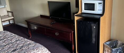 A hotel room with a bed, a dresser, a television, a microwave, a trash bin, and a hanging light fixture.