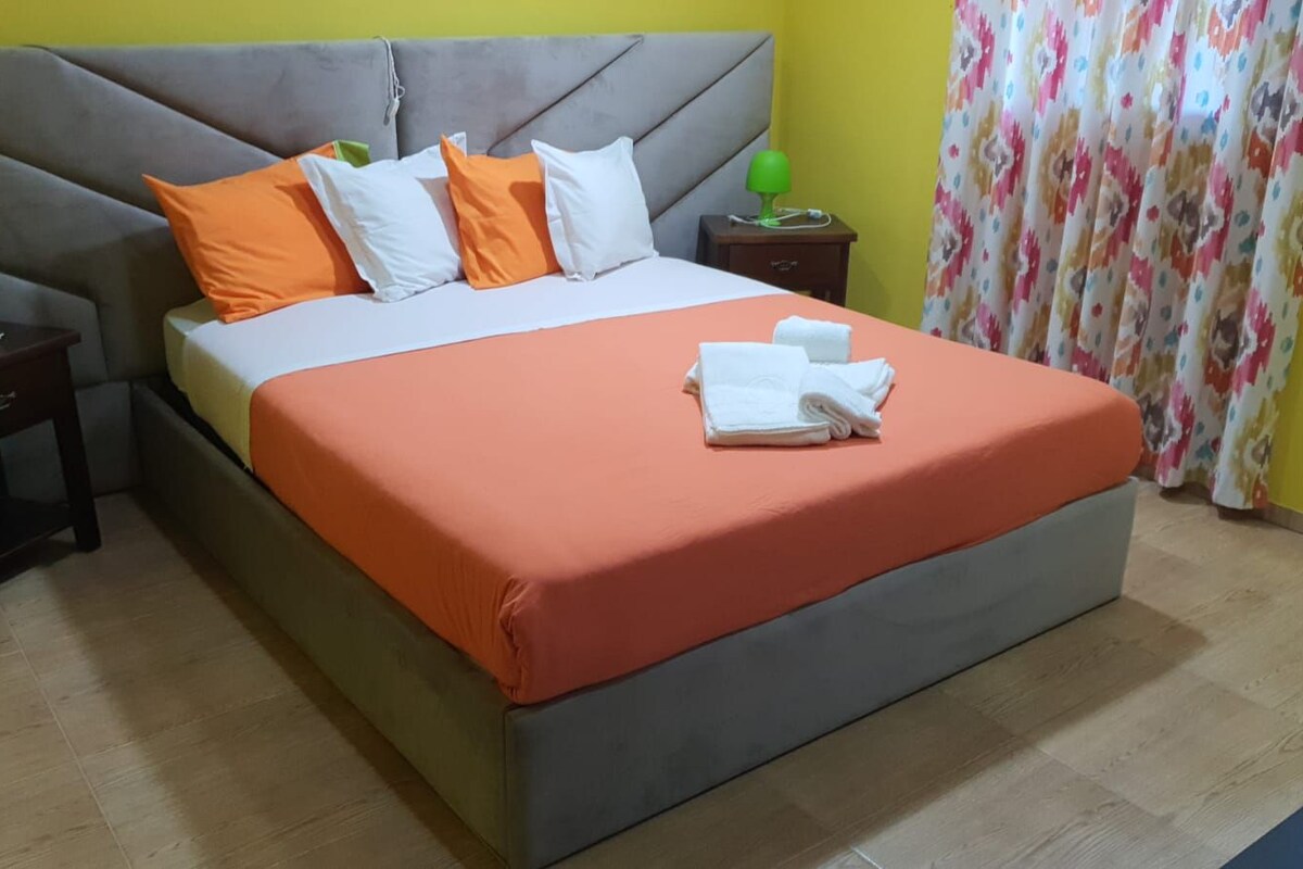 Una cama con cabecera gris, ropa de cama naranja y blanca, una mesita de noche verde y una cortina con estampado floral.