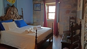 Une chambre à coucher avec un lit, une tête de lit en bois, un mur en pierre, une fenêtre avec des rideaux et une petite table.