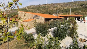 Un bâtiment à la façade en pierre et au toit recouvert de tuiles rouges, entouré de verdure et adossé à une colline où l’on aperçoit des éoliennes au loin.