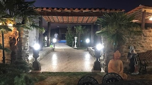 Une cour avec un sol carrelé, des palmiers et un mur en pierre. On y trouve deux statues et une pergola illuminée.