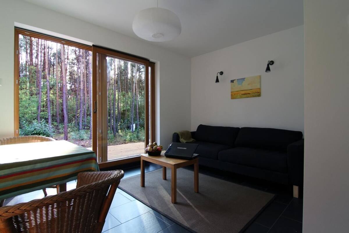 A modern living room with a large sliding glass door leading to a forest view, a black sofa, a small table with a laptop, and a wicker chair.