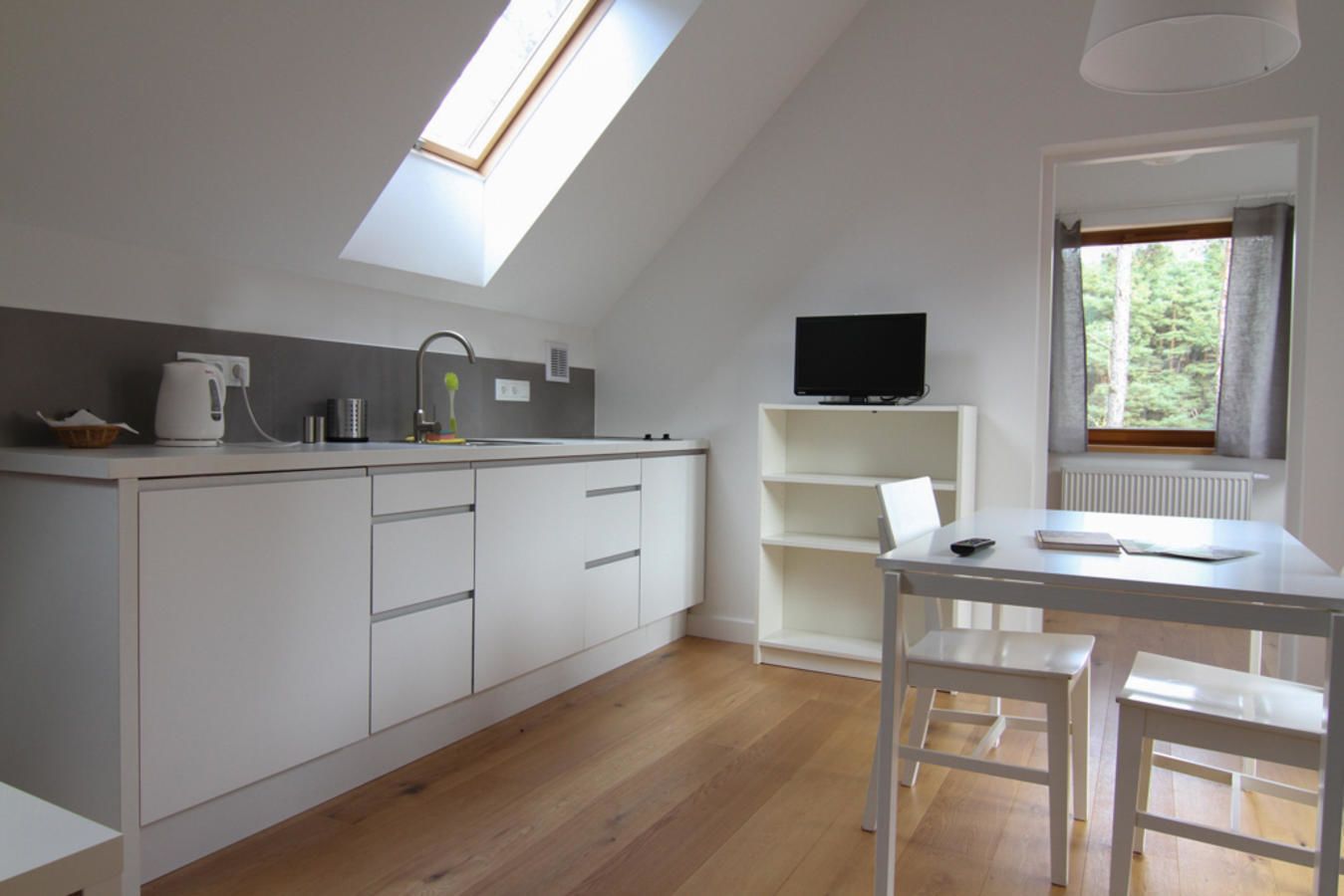 A modern kitchen with a skylight, white cabinetry, a dining area with a table and chairs, and a TV on a shelf.