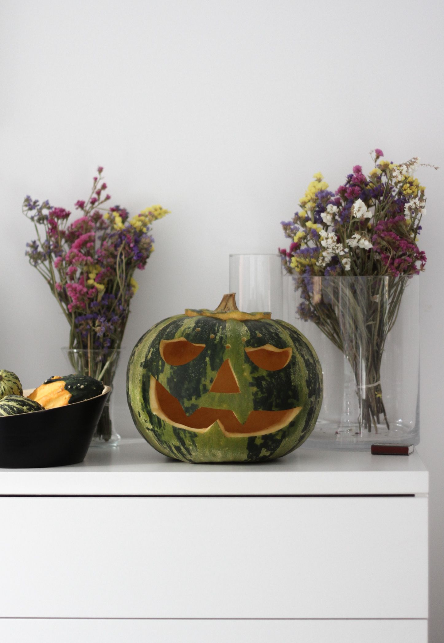 Una calabaza tallada con rostro sobre una superficie blanca, flanqueada por dos jarrones con flores.