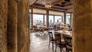Um restaurante com paredes de pedra, vigas de madeira e grandes janelas que oferecem vista para a cidade. Mesas com cadeiras e toalhas.