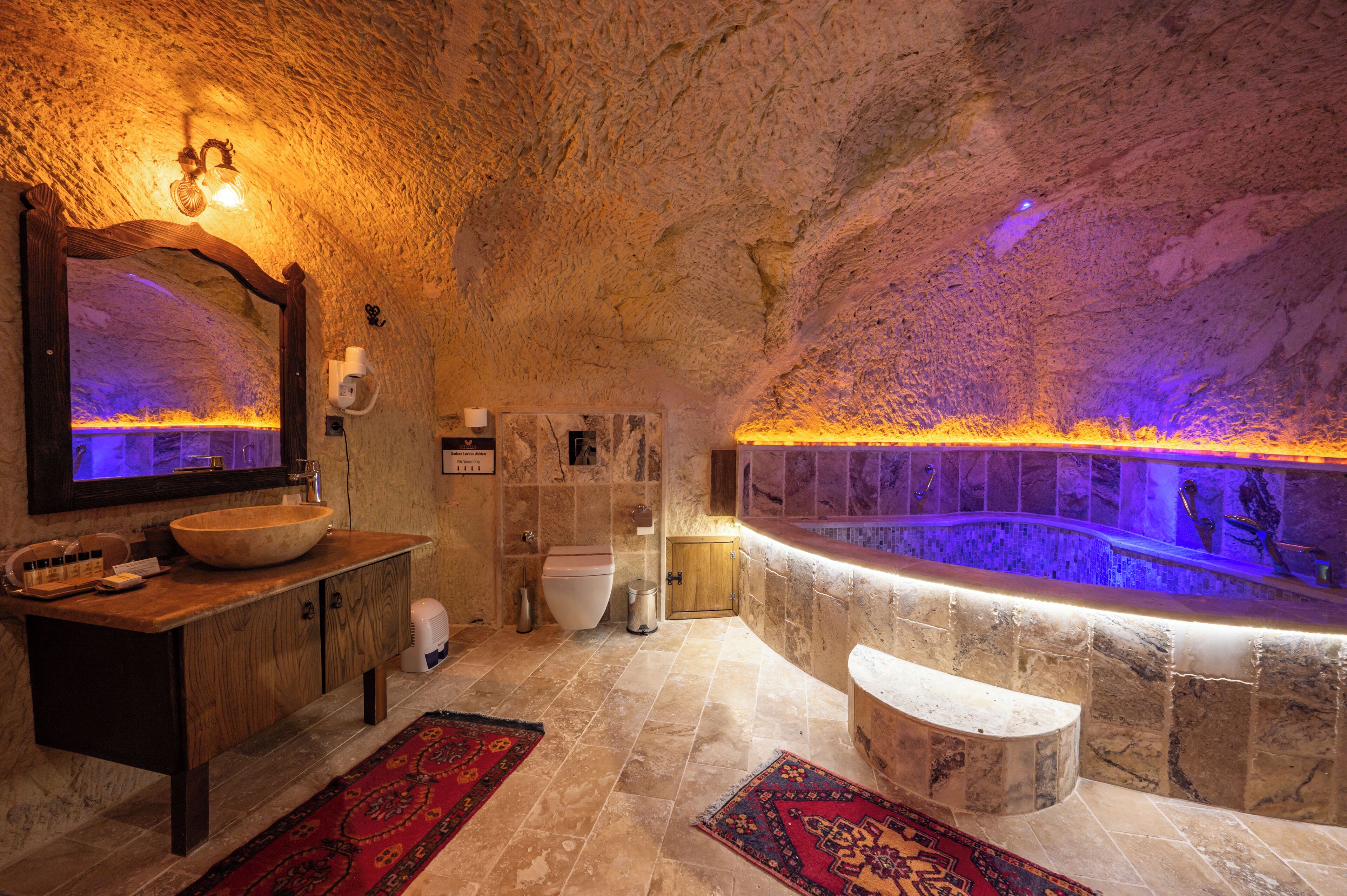 A spa-like bathroom with a stone wall, a wooden vanity, a large mirror, a bathtub, and a red patterned rug.