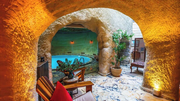 Arco de pedra que dá acesso a uma área de piscina, com um banco de madeira e uma planta em vaso.