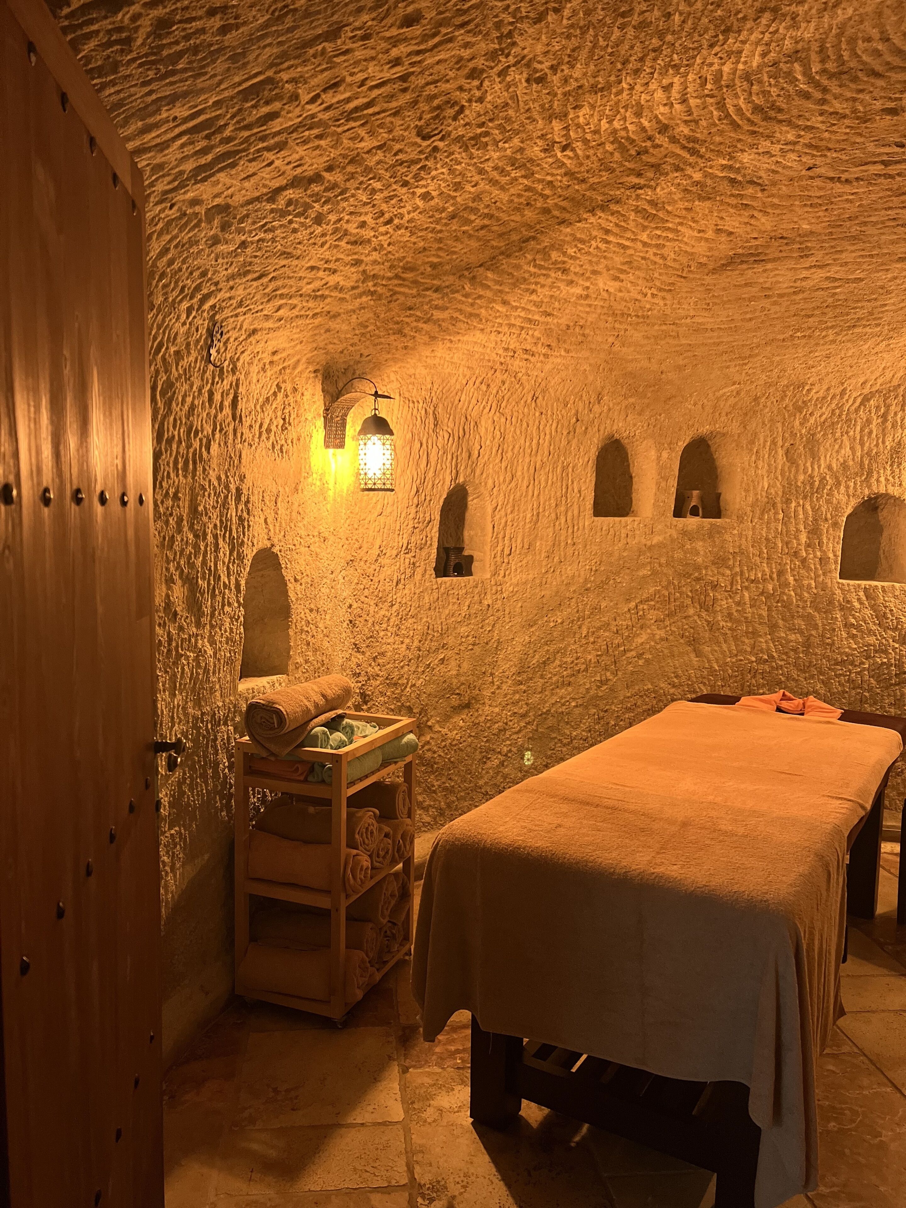 Ein Zimmer mit Steindecke, einer Holztür, einem Bett mit weißer Decke, einer Holzbank mit Handtüchern und einer Wandleuchte.