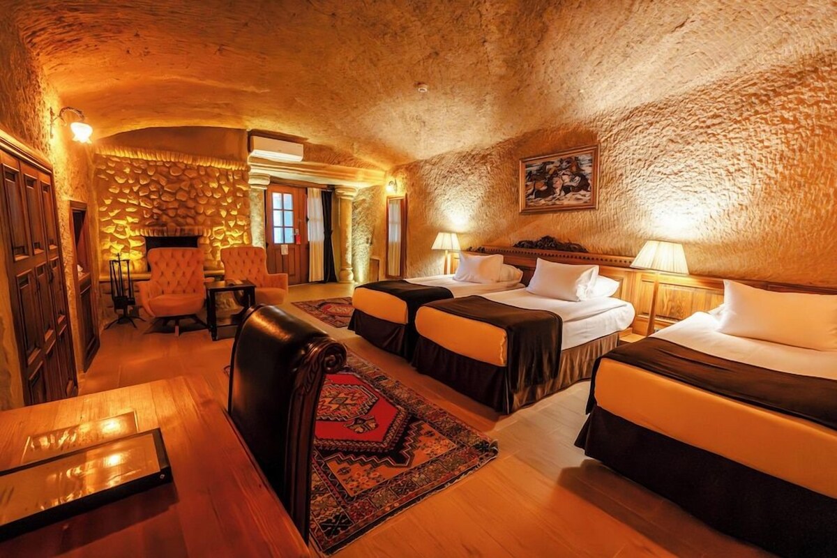 Um quarto com teto de pedra, duas camas com lençóis brancos, uma mesa de madeira e um tapete estampado.