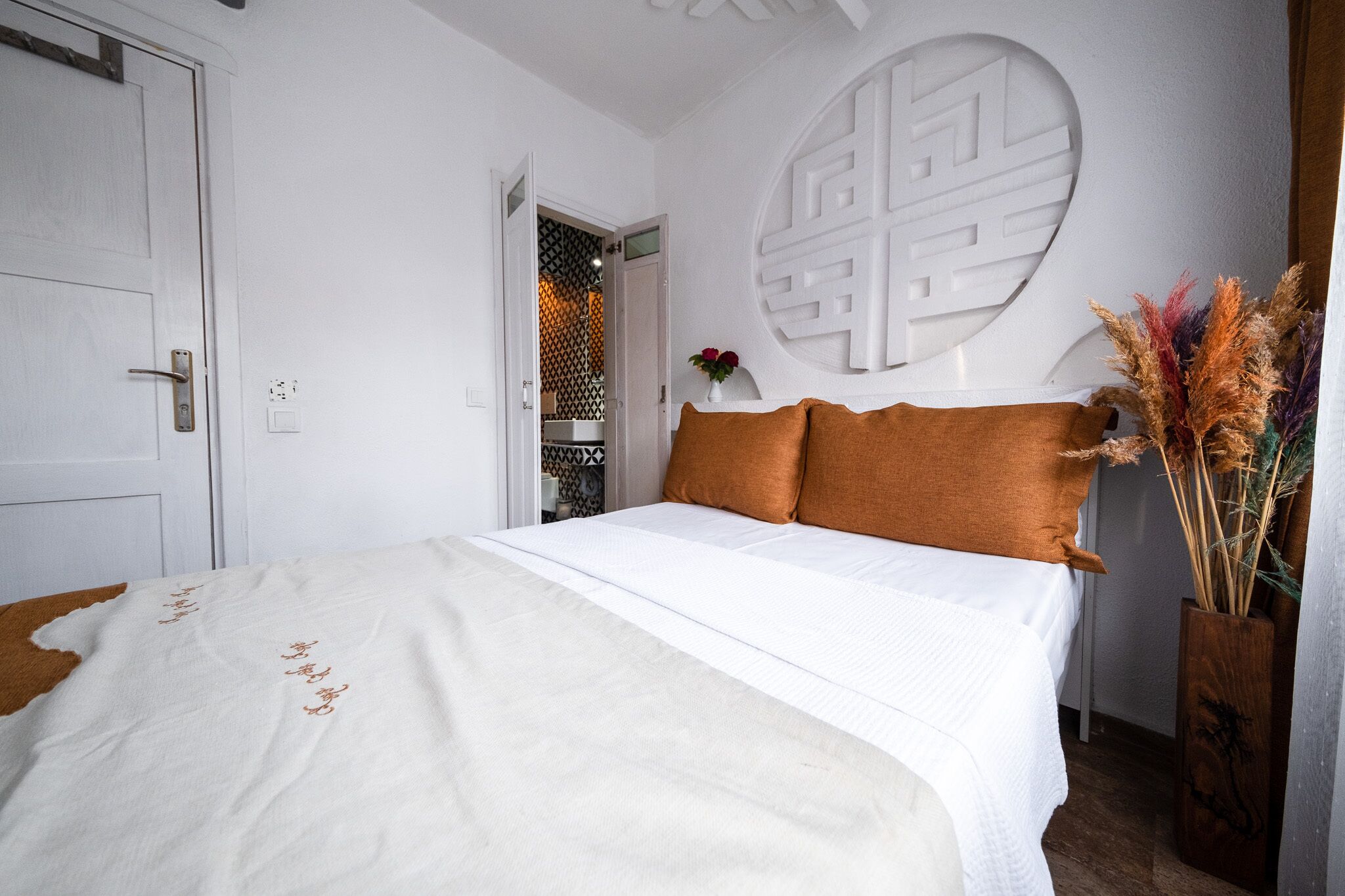 A bedroom with a bed, white walls, a decorative wall piece, and a vase with dried plants.