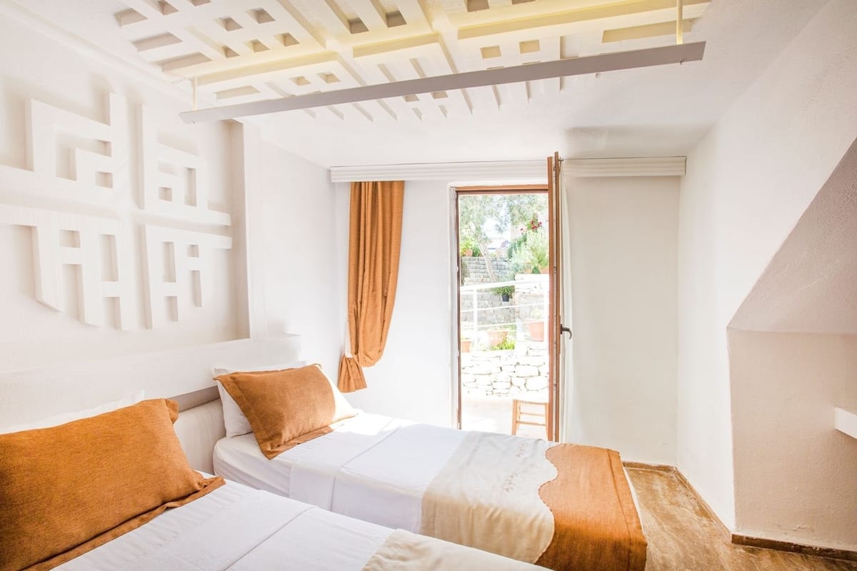 A bedroom with two beds, a wooden ceiling, and a view of a garden through a sliding door.