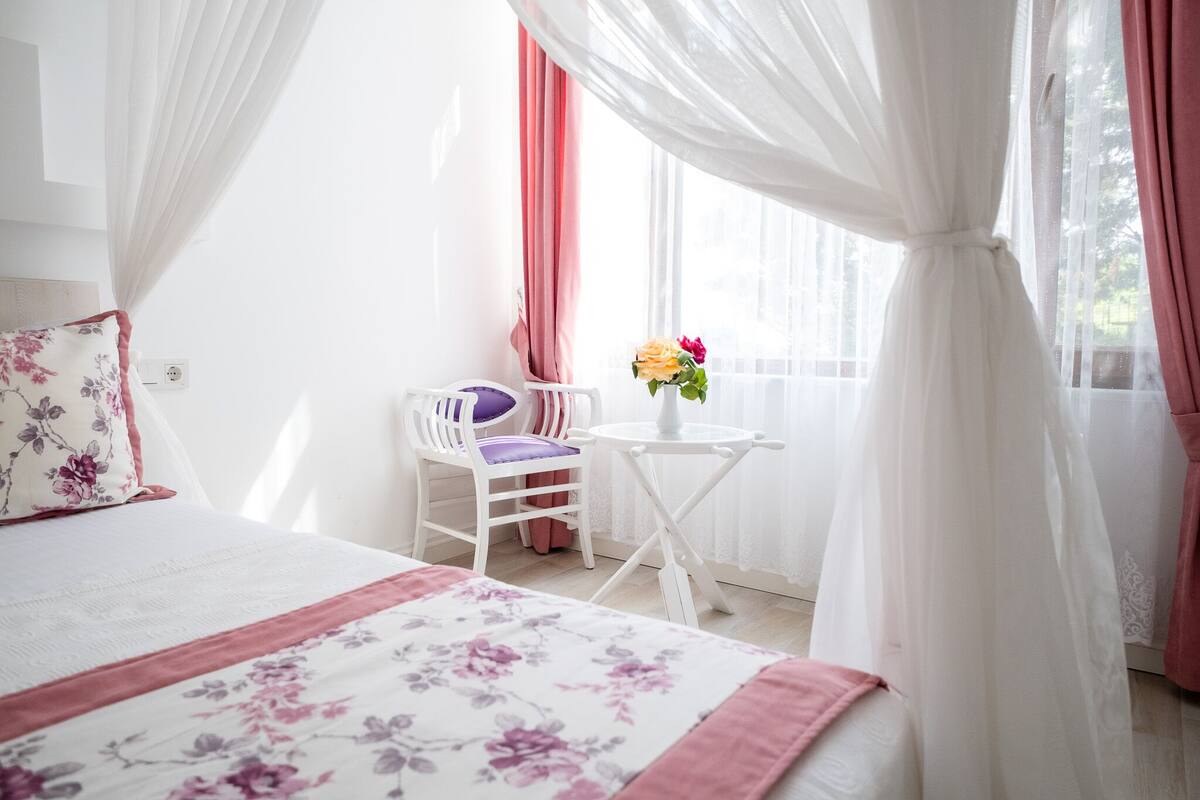 A bedroom with a bed, a chair, a small table with flowers, and sheer curtains.